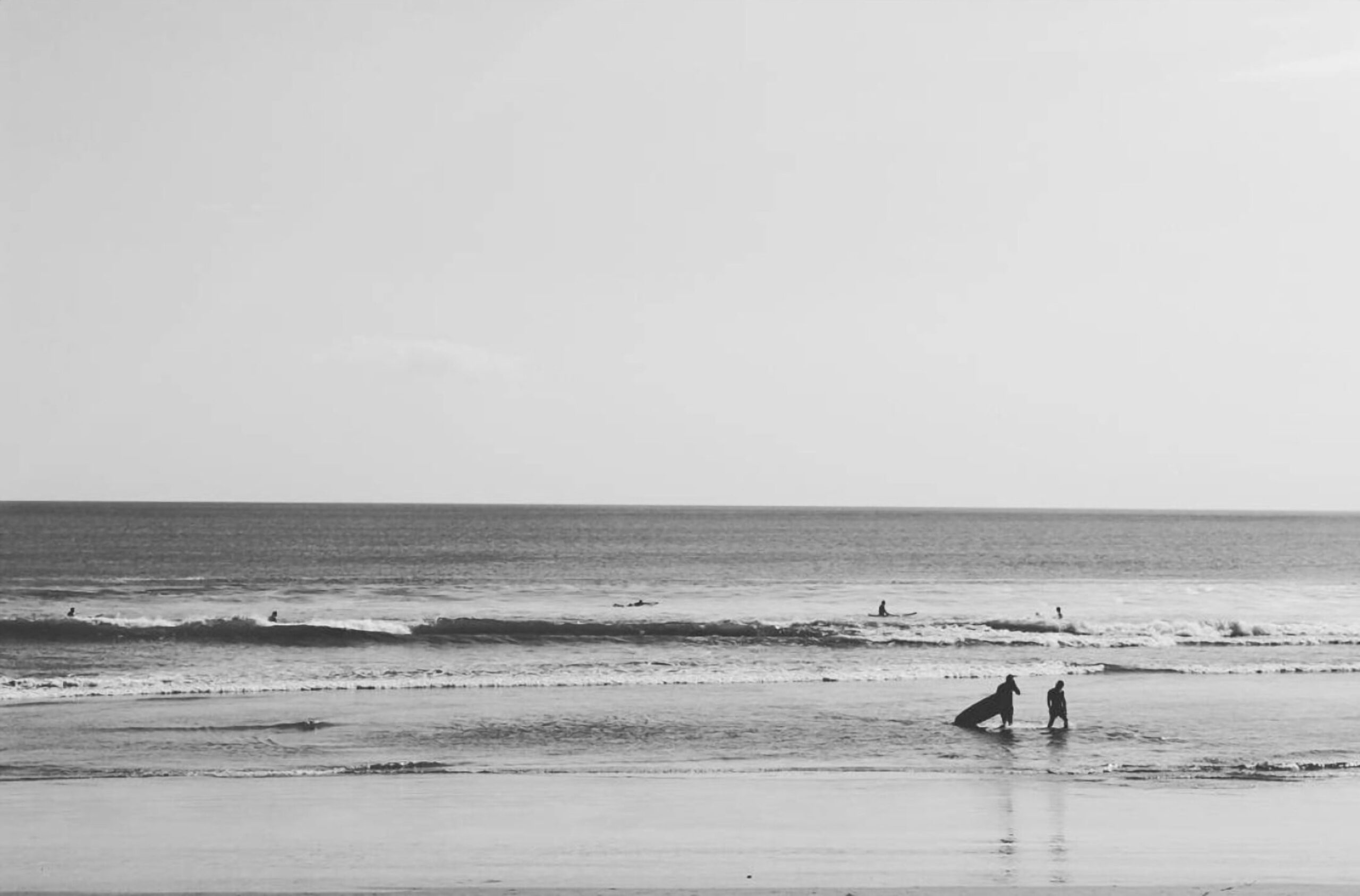 Two surfers walking out of the sea on a wide beach, black and white